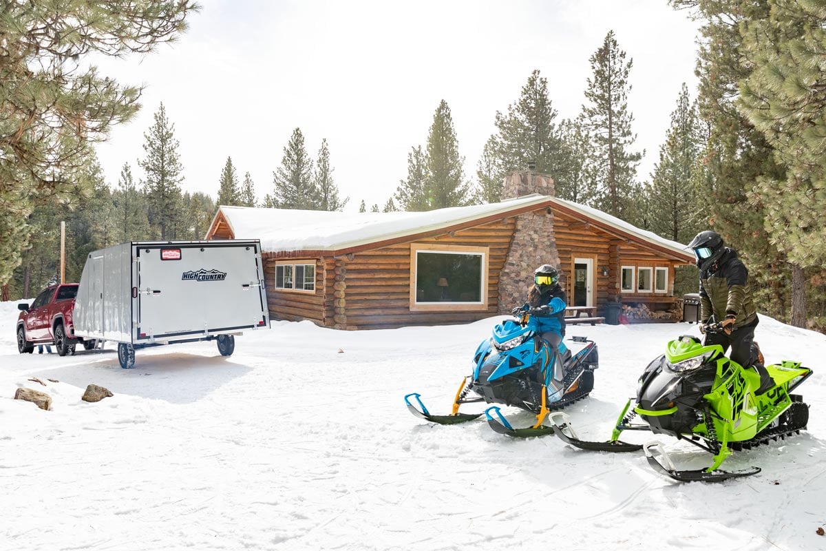 Crossover aluminum snowmobile trailer parked at a cabin with two sleds ready to ride.