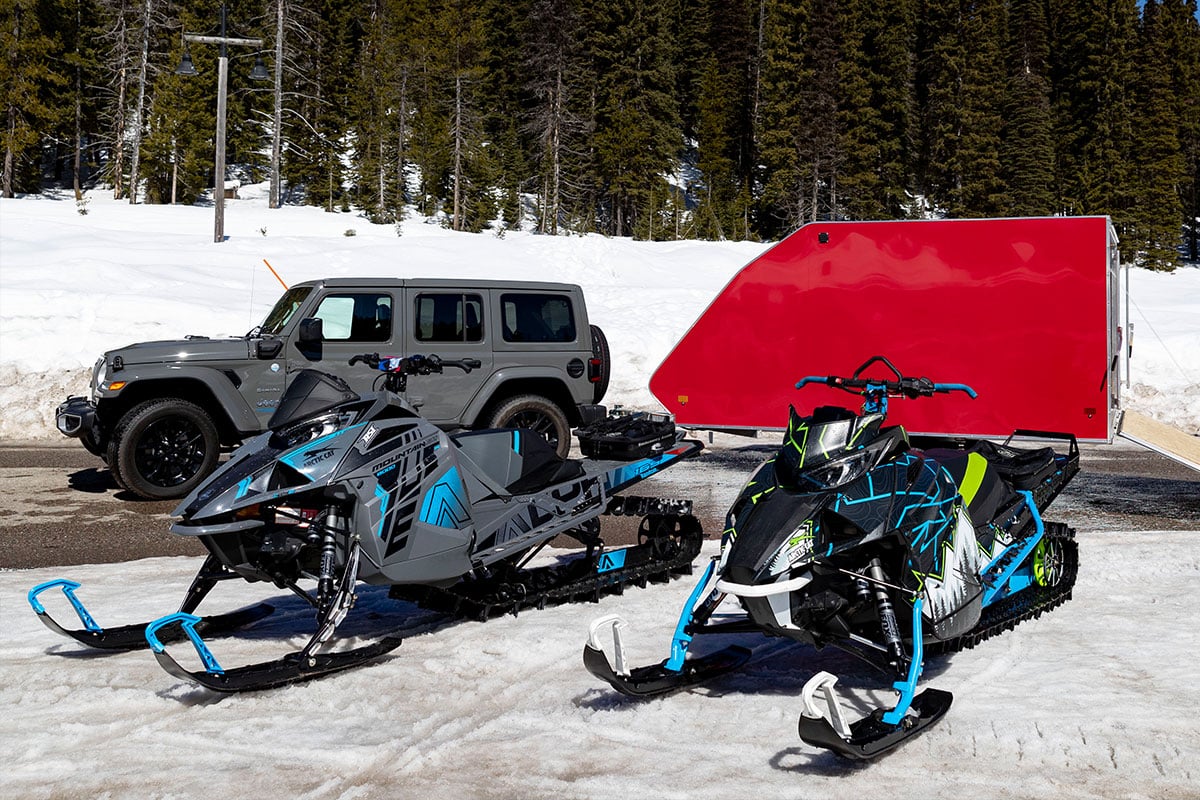 A red enclosed ALCOM crossover/hybrid snow trailer and two snowmobiles parked in front of it on a snowy day.