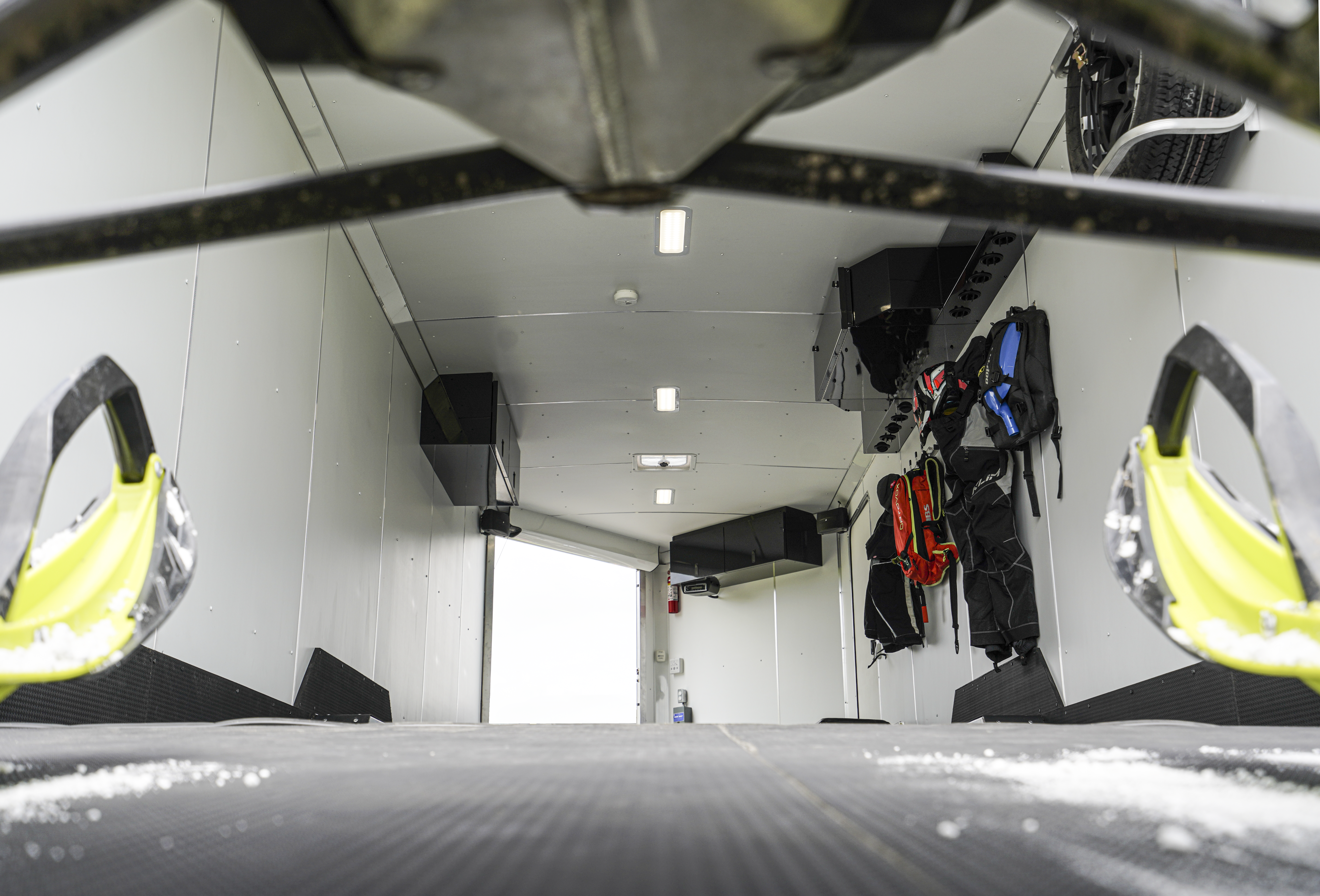 View from between snowmobile skis inside a well maintained and decked out aluminum snowmobile trailer with cabinets, lights, doors and coat hooks