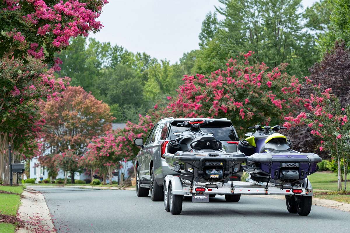 Silver SUV towing a Triton aluminum two-place PWC trailer through a scenic neighborhood with pink flowering trees.