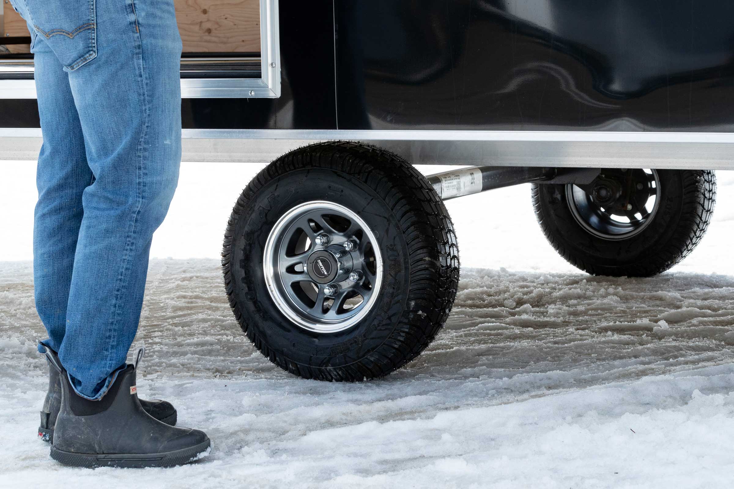 Close view of tires on an ALCOM aluminum snowmobile trailer and a man's feet standing beside it
