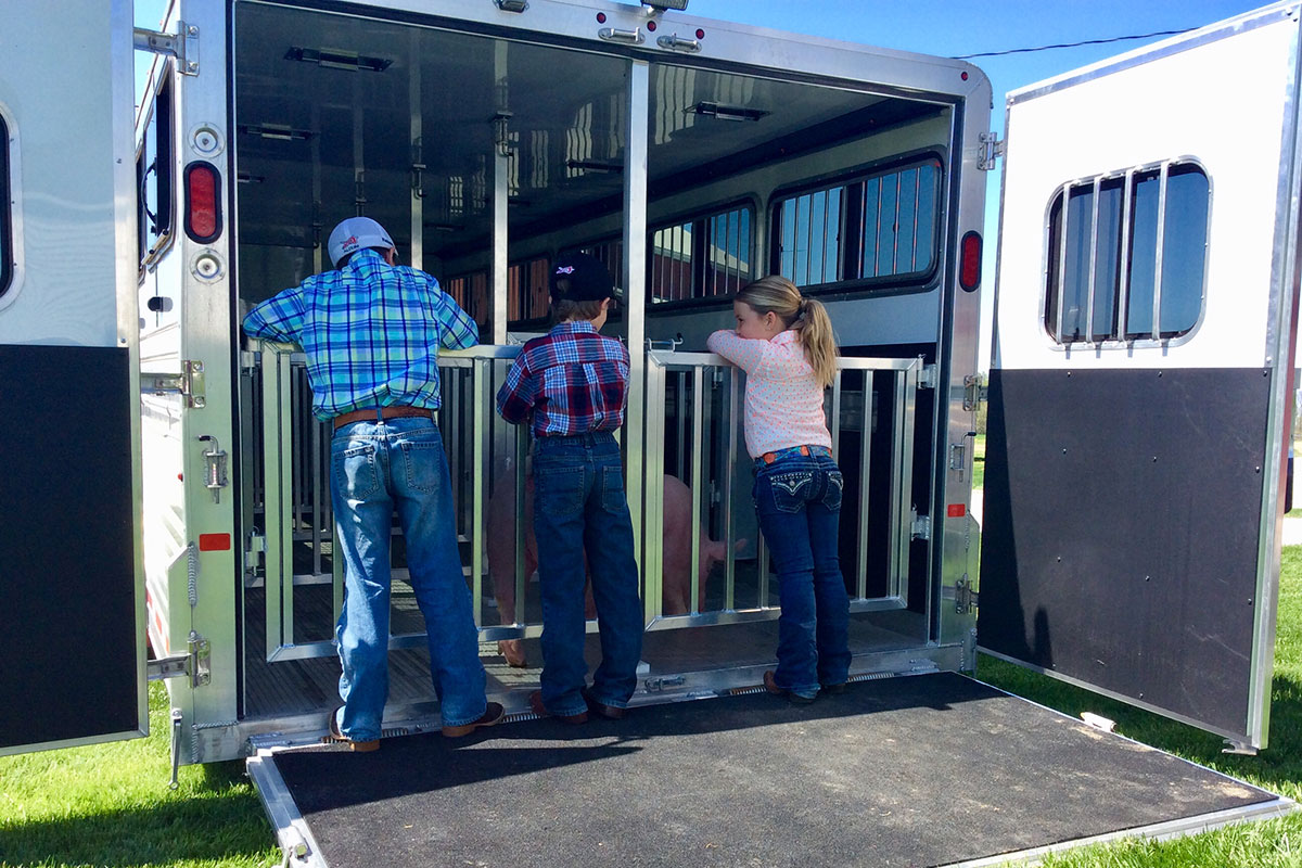 Three young farmers in training stand at the open loading end of a Frontier Stock LowPro trailer with pigs loaded safely into adjustable stalls.