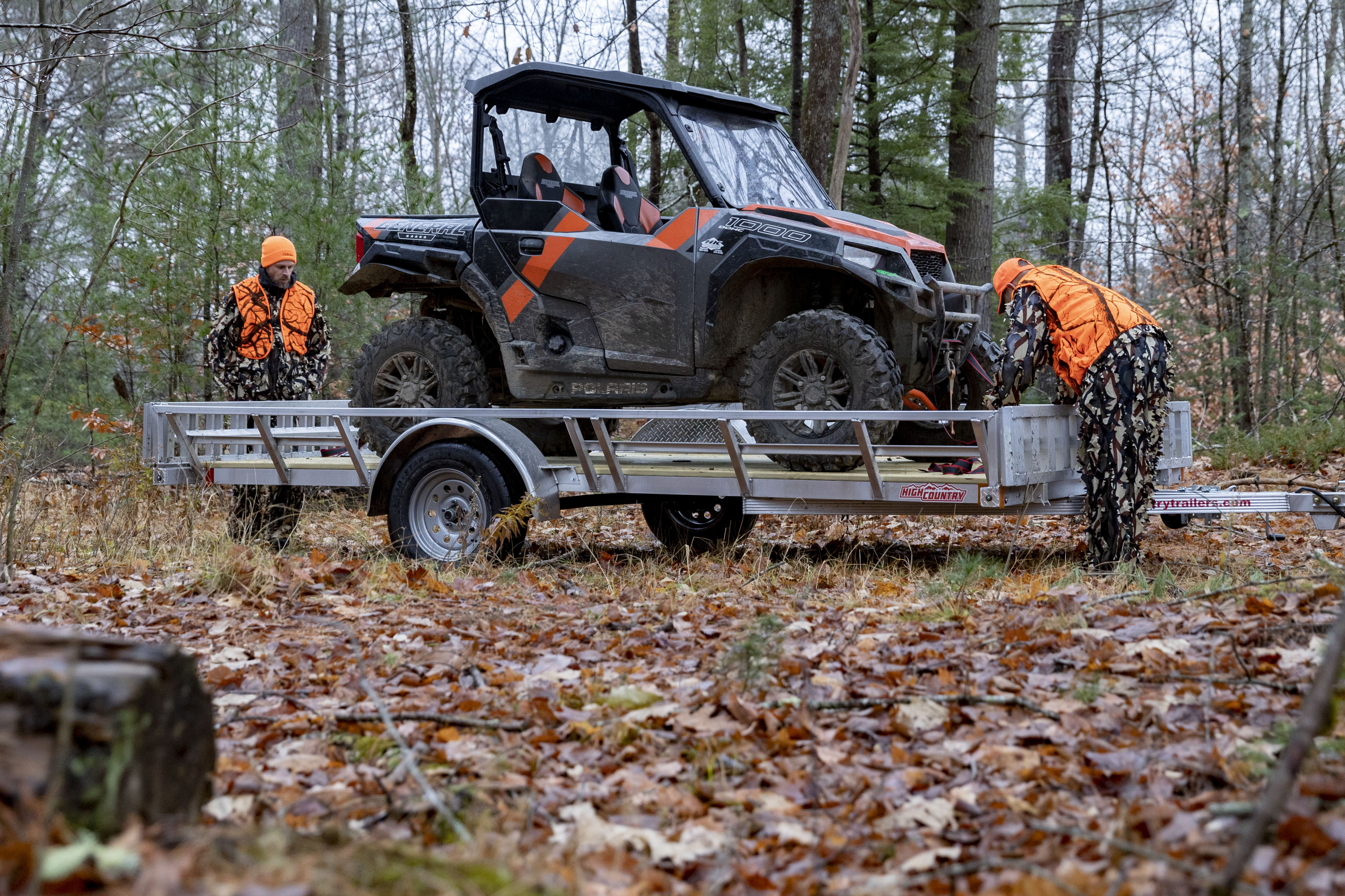 Two men in camo suits and safety orange vests and hats prepare to unload a UTV from an aluminum UTV trailer parked on a trail during autumn